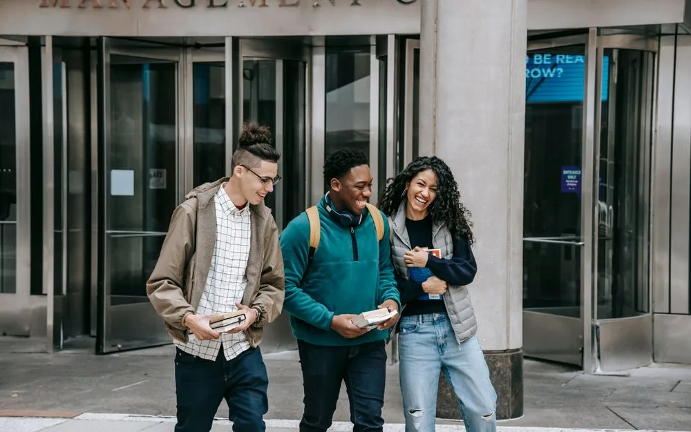 Three university friends carrying books and laughing outside a campus building